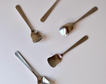 May include: A set of five silver-toned metal spoons with a polished finish. Each spoon features a long, slender handle and a wide, flat, square-shaped bowl with slightly curved edges, arranged in a scattered pattern on a plain surface.