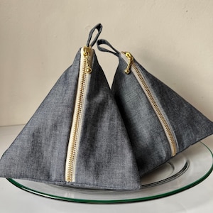 May include: Two denim-like triangular pouches with gold-colored zippers and small loop handles. The pouches are displayed on a clear glass plate. The zippers run down the center of the pouches.