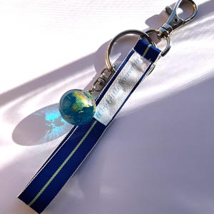 Puede incluir: Un llavero con un dije de globo terráqueo y una correa a rayas azules y blancas. El globo muestra un mapa detallado del mundo. La correa tiene el texto "by Little Souvenir, New York". Incluye un mosquetón y un llavero plateados.