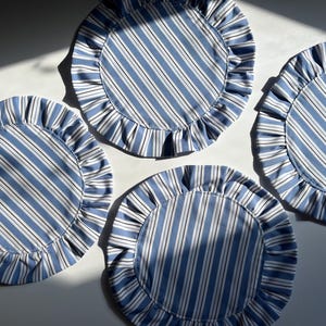 May include: Four circular placemats featuring a blue and white striped design, each with a ruffled border. The placemats are arranged on a white surface, with shadows adding depth to the image.
