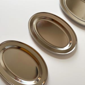 以下が含まれることがあります： 白い表面に並んだ、縁が盛り上がった銀色の楕円形のサービングディッシュが3つあります。