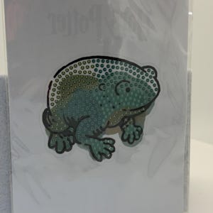 May include: A craft kit featuring a green frog design made of small, colorful diamond-shaped pieces. The frog is on a white background, and the kit is sealed in clear plastic. The word "Potter" is visible in the background.