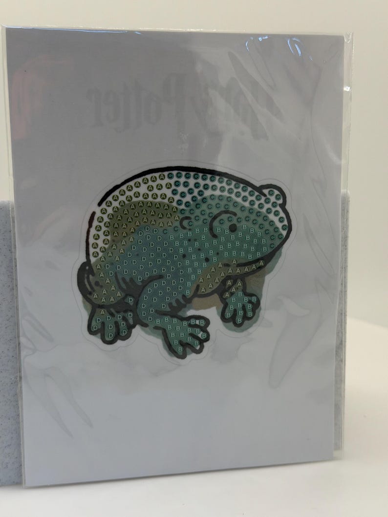 May include: A craft kit featuring a green frog design made of small, colorful diamond-shaped pieces. The frog is on a white background, and the kit is sealed in clear plastic. The word "Potter" is visible in the background.