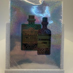 May include: A holographic sticker featuring two potion bottle designs. The left bottle reads "Dragon Blood" and the right bottle reads "Acromantula Venom". The sticker is displayed in a gray felt stand.