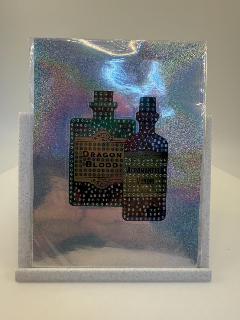 May include: A holographic sticker featuring two potion bottle designs. The left bottle reads "Dragon Blood" and the right bottle reads "Acromantula Venom". The sticker is displayed in a gray felt stand.