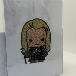 May include: A diamond painting kit featuring a cartoon character with blonde hair and a black outfit. The image is on a white card, encased in clear plastic. The character is holding a staff.