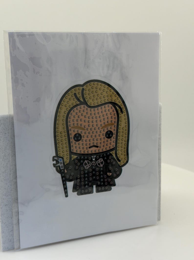 May include: A diamond painting kit featuring a cartoon character with blonde hair and a black outfit. The image is on a white card, encased in clear plastic. The character is holding a staff.