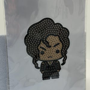 May include: A sticker featuring a stylized illustration of a person with dark hair and a black outfit. The design uses a pattern of letters, including "B", "A", and "C", to create the image. The sticker is in a clear plastic sleeve.