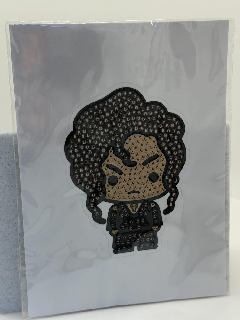 May include: A sticker featuring a stylized illustration of a person with dark hair and a black outfit. The design uses a pattern of letters, including "B", "A", and "C", to create the image. The sticker is in a clear plastic sleeve.