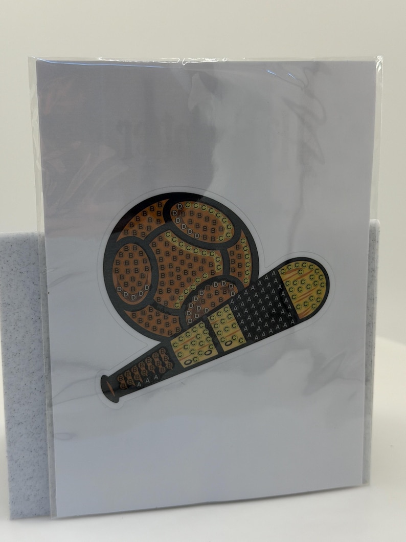 Puede incluir: Una pegatina con un dise&ntilde;o de b&eacute;isbol y bate. La pelota de b&eacute;isbol es marr&oacute;n con un patr&oacute;n de peque&ntilde;as formas amarillas y marrones. El bate es negro y amarillo con un patr&oacute;n texturizado. La pegatina est&aacute; en una tarjeta blanca.