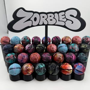 May include: A display of colorful, spherical toys, each with a unique pattern and color scheme, arranged in a black tiered holder. A black sign with the word "ZORBLES" in white lettering sits atop the display.