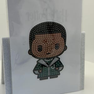 Puede incluir: Una pegatina con un personaje de dibujos animados con un dise&ntilde;o de mosaico. El personaje tiene una tez oscura y viste un atuendo verde y negro. El mosaico est&aacute; formado por las letras A, B y D. La pegatina se muestra en una tarjeta blanca.