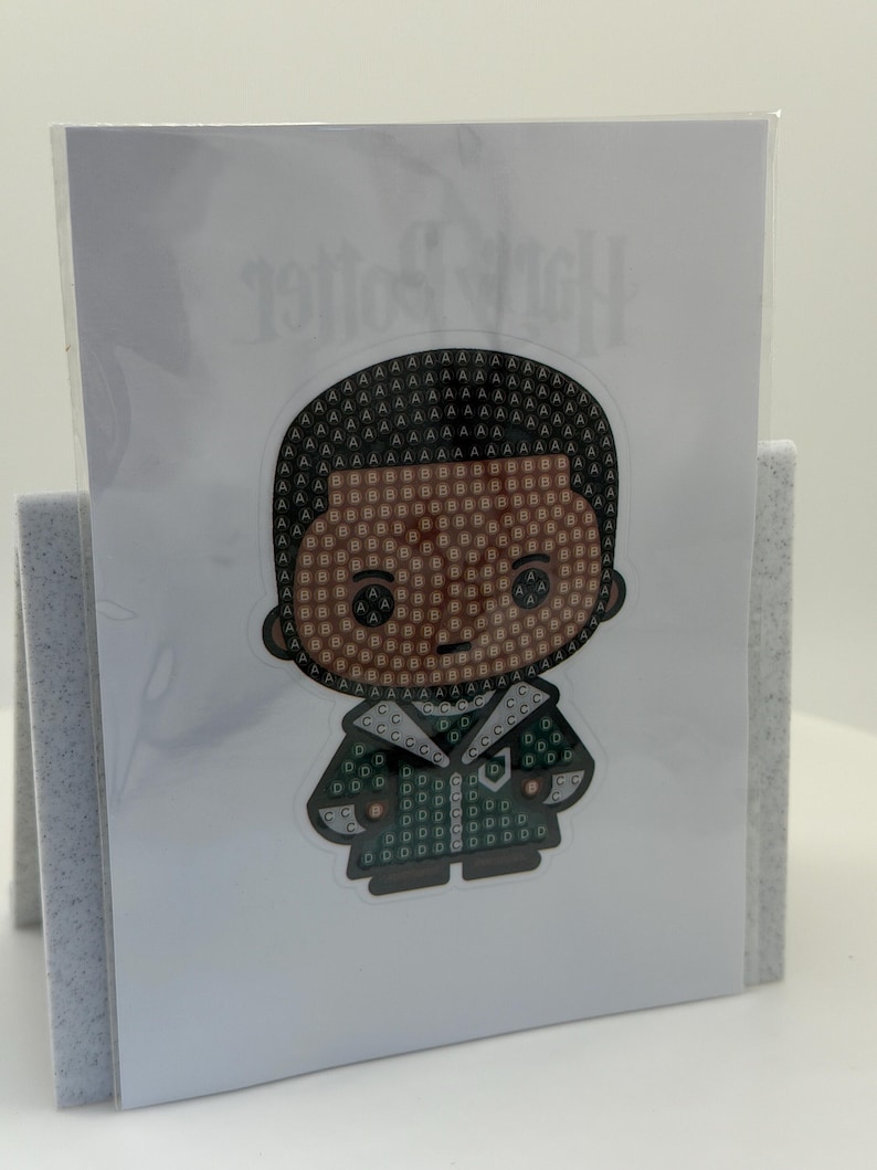 Puede incluir: Una pegatina con un personaje de dibujos animados con un dise&ntilde;o de mosaico. El personaje tiene una tez oscura y viste un atuendo verde y negro. El mosaico est&aacute; formado por las letras A, B y D. La pegatina se muestra en una tarjeta blanca.