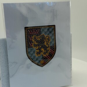 Puede incluir: Un emblema en forma de escudo con un le&oacute;n dorado rampante sobre un fondo estampado de color rojo, verde y azul. El emblema tiene un borde dorado y est&aacute; en una funda de pl&aacute;stico transparente, presentado en una tarjeta blanca.