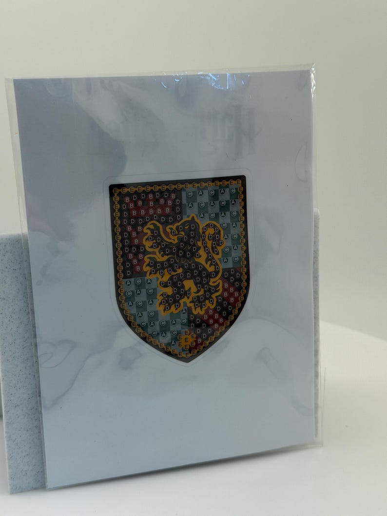 Puede incluir: Un emblema en forma de escudo con un le&oacute;n dorado rampante sobre un fondo estampado de color rojo, verde y azul. El emblema tiene un borde dorado y est&aacute; en una funda de pl&aacute;stico transparente, presentado en una tarjeta blanca.