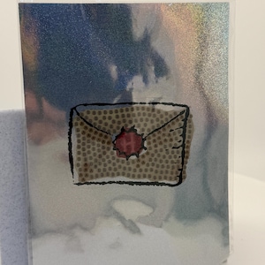 May include: A holographic card featuring a brown envelope design with a red wax seal. The envelope is covered in a pattern of the letter "B". The card is encased in a clear plastic sleeve, with a blue and silver background.