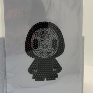 May include: A black and white sticker featuring a stylized figure with a rounded head and a cloak-like body. The design is composed of letters and swirls, creating a unique pattern. The sticker is encased in a clear plastic sleeve.