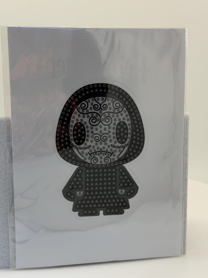 May include: A black and white sticker featuring a stylized figure with a rounded head and a cloak-like body. The design is composed of letters and swirls, creating a unique pattern. The sticker is encased in a clear plastic sleeve.