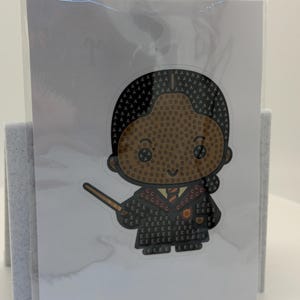Puede incluir: Una pegatina con un personaje de dibujos animados con piel morena, pelo negro y una varita m&aacute;gica. El personaje lleva una t&uacute;nica negra con un emblema dorado y rojo. La pegatina est&aacute; sobre una tarjeta blanca, sujeta en un soporte de fieltro gris.