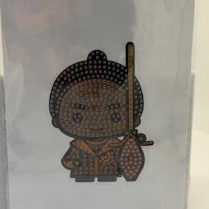 Puede incluir: Una pegatina con un personaje de dibujos animados con cabeza marr&oacute;n y atuendo marr&oacute;n. El personaje sostiene una espada amarilla y negra. La pegatina est&aacute; cubierta de peque&ntilde;as letras y s&iacute;mbolos.