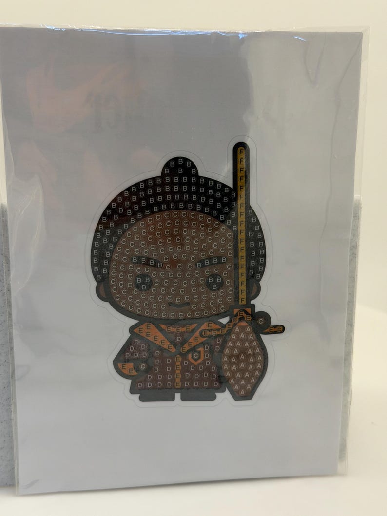 Puede incluir: Una pegatina con un personaje de dibujos animados con cabeza marr&oacute;n y atuendo marr&oacute;n. El personaje sostiene una espada amarilla y negra. La pegatina est&aacute; cubierta de peque&ntilde;as letras y s&iacute;mbolos.