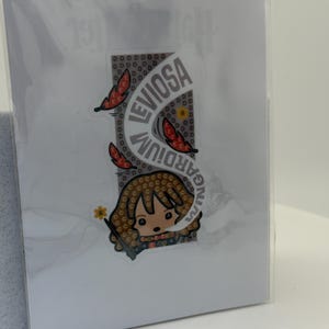May include: A sticker featuring a cartoon character with brown hair and a wand, with the words "LEVIOSA" and "WINGARDIUM" in a curved design. The sticker is on a white background, partially obscured by plastic.