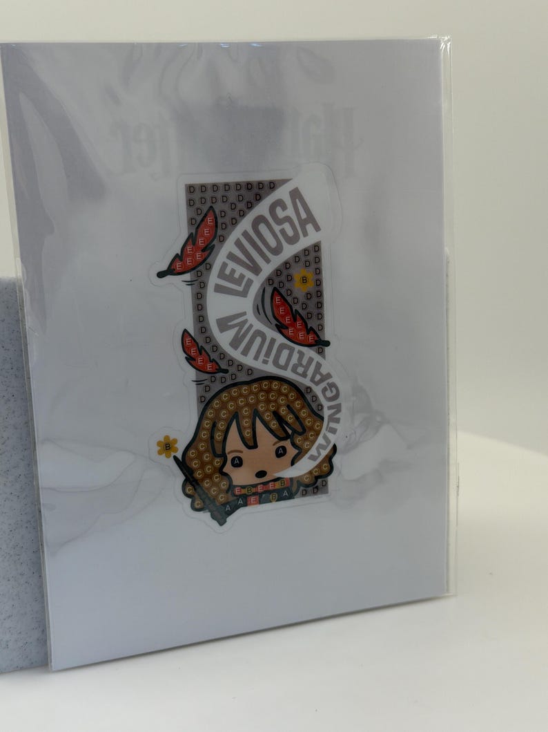 May include: A sticker featuring a cartoon character with brown hair and a wand, with the words "LEVIOSA" and "WINGARDIUM" in a curved design. The sticker is on a white background, partially obscured by plastic.