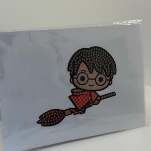 Puede incluir: Un kit de pintura de diamantes con una imagen de dibujos animados de un personaje montado en una escoba. El personaje tiene pelo negro, gafas y un atuendo rojo y dorado. La imagen est&aacute; sobre un fondo blanco y en una funda de pl&aacute;stico transparente.