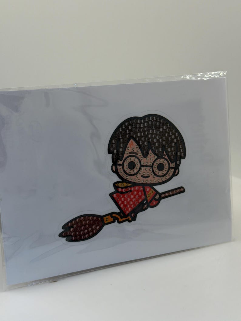 Puede incluir: Un kit de pintura de diamantes con una imagen de dibujos animados de un personaje montado en una escoba. El personaje tiene pelo negro, gafas y un atuendo rojo y dorado. La imagen est&aacute; sobre un fondo blanco y en una funda de pl&aacute;stico transparente.