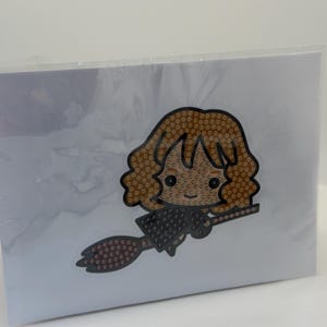 Puede incluir: Un kit de manualidades empaquetado con un personaje de dibujos animados con cabello casta&ntilde;o y una escoba. El personaje est&aacute; hecho de peque&ntilde;as cuentas de colores. El fondo es blanco.