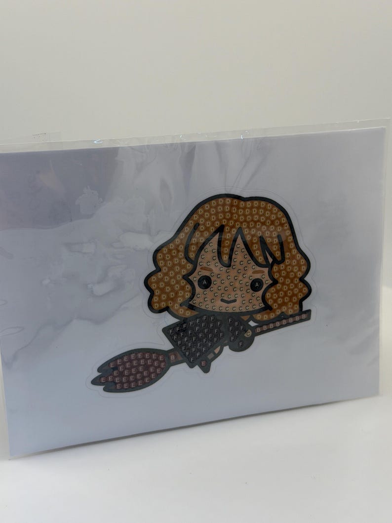 Puede incluir: Un kit de manualidades empaquetado con un personaje de dibujos animados con cabello casta&ntilde;o y una escoba. El personaje est&aacute; hecho de peque&ntilde;as cuentas de colores. El fondo es blanco.
