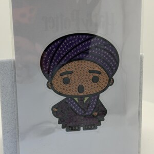 May include: A sticker featuring a cartoon character in a purple robe and hat. The character's face is tan with brown eyes and a small smile. The sticker is on a white background and is in a clear plastic sleeve.