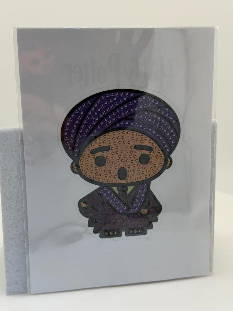 May include: A sticker featuring a cartoon character in a purple robe and hat. The character's face is tan with brown eyes and a small smile. The sticker is on a white background and is in a clear plastic sleeve.