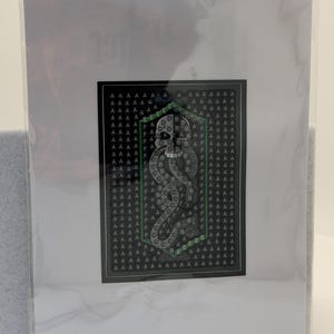 Puede incluir: Una impresi&oacute;n art&iacute;stica enmarcada con un fondo negro y un patr&oacute;n repetido de la letra "A". El emblema central, con borde verde, muestra una serpiente estilizada y las letras "B", "C", "D", "E" y "F".