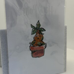 May include: A clear sticker featuring a cartoon mandrake plant in a terracotta pot. The mandrake has a green leafy head and a brown body. The pot is orange with a repeating pattern. The sticker is on a white card.