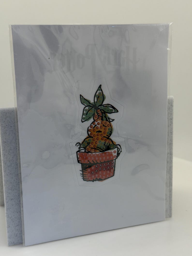 May include: A clear sticker featuring a cartoon mandrake plant in a terracotta pot. The mandrake has a green leafy head and a brown body. The pot is orange with a repeating pattern. The sticker is on a white card.