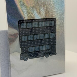 Puede incluir: Una tarjeta de felicitaci&oacute;n con un dise&ntilde;o de autob&uacute;s, la palabra "HELLO" est&aacute; formada por letras y formas repetidas. El autob&uacute;s es negro y gris, sobre un fondo plateado hologr&aacute;fico. La tarjeta est&aacute; dentro de una funda de pl&aacute;stico transparente.