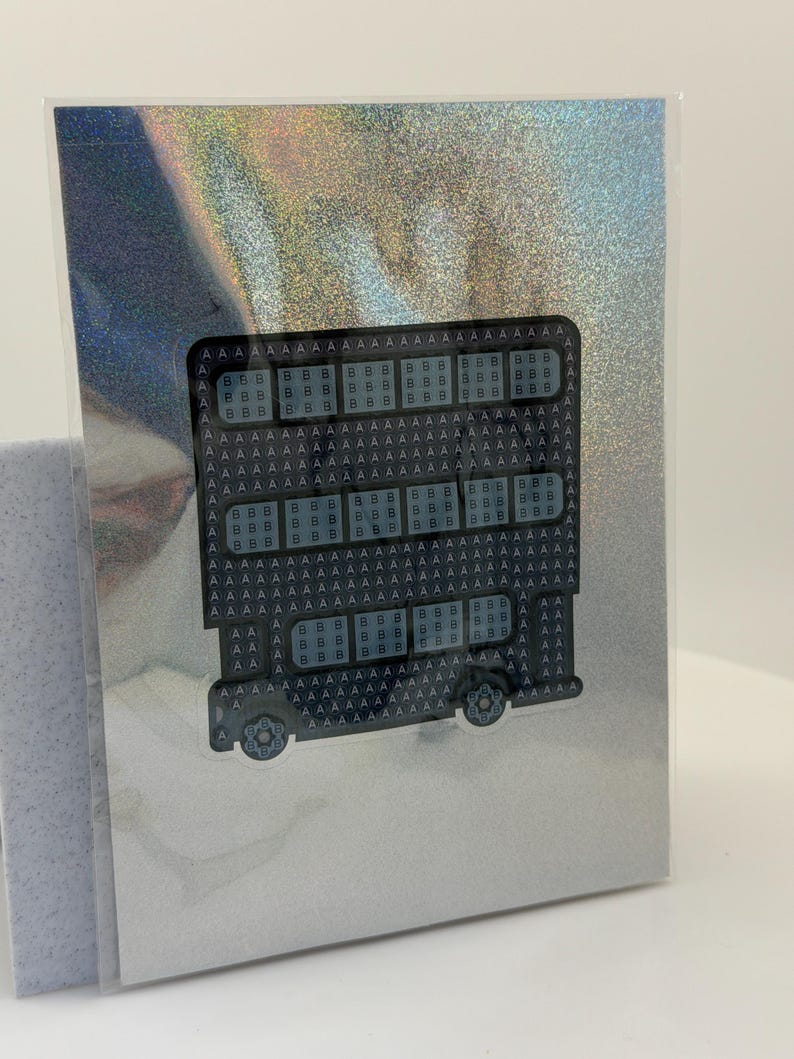 Puede incluir: Una tarjeta de felicitaci&oacute;n con un dise&ntilde;o de autob&uacute;s, la palabra "HELLO" est&aacute; formada por letras y formas repetidas. El autob&uacute;s es negro y gris, sobre un fondo plateado hologr&aacute;fico. La tarjeta est&aacute; dentro de una funda de pl&aacute;stico transparente.
