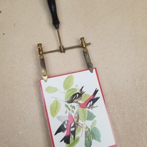 May include: A brass picture frame with a wooden handle and two adjustable arms. The frame is holding a card with a colorful illustration of three birds on a branch.