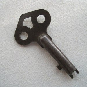 May include: A vintage metal key with a unique, three-hole design in the head. The key is made of dark metal and has a long, narrow shaft.