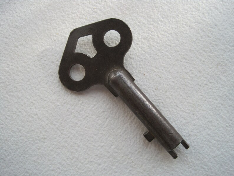 May include: A vintage metal key with a unique, three-hole design in the head. The key is made of dark metal and has a long, narrow shaft.