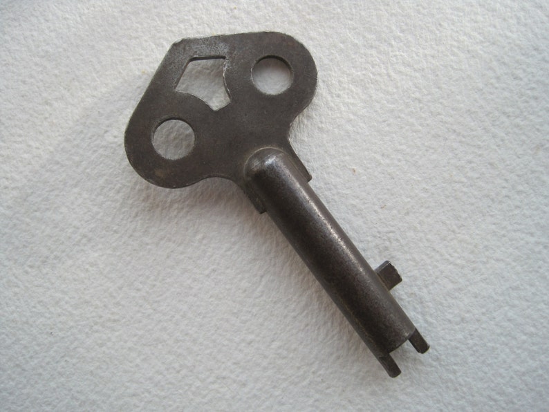 May include: A vintage metal key with a unique, irregular shaped head featuring three holes. The key is made of a dark, aged metal and has a long, straight shaft.
