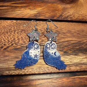 May include: A pair of blue glitter owl earrings with a silver star charm. The owls feature a moon and stars design.