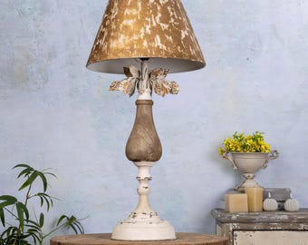 May include: A white and brown table lamp with a gold leaf design on the base and a gold and white patterned lampshade. The lamp is sitting on a wooden table.