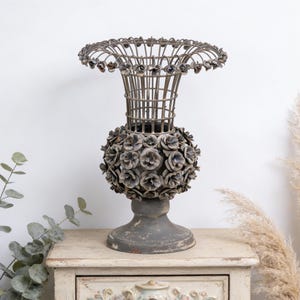 May include: An antique-style metal vase with a distressed gray finish. The vase features a spherical base adorned with floral accents and a cage-like upper section. The vase is displayed on a vintage-style wooden cabinet.