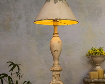 May include: A distressed, cream-coloured table lamp with a conical lampshade. The shade has a faded, aged appearance with a yellow interior. The lamp's base is ornate and painted to look aged. The lamp is lit, casting a warm glow.