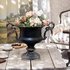 May include: A black metal vase with ornate handles sits on a wooden table. The vase is filled with pink and white flowers.