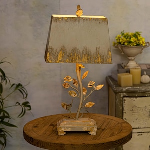 Peut inclure: Une lampe de table blanche et dorée vieillie avec un motif floral. La lampe a un abat-jour carré et une base avec un motif floral.