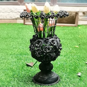 Op de afbeelding: Een zwarte metalen vaas met een rozenontwerp, met roze en witte tulpen. De vaas heeft een ronde basis en een lang, slank lichaam.