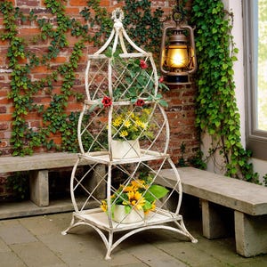 May include: White metal plant stand with three tiers and a decorative top. The stand is decorated with a diamond pattern and has a distressed finish. The stand is filled with potted plants, including sunflowers and red roses.